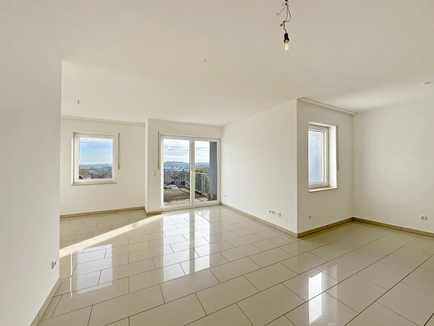 Bright, empty room with white walls and beige tile floor. Windows and sliding glass doors offer city views. A bare bulb hangs from the ceiling.