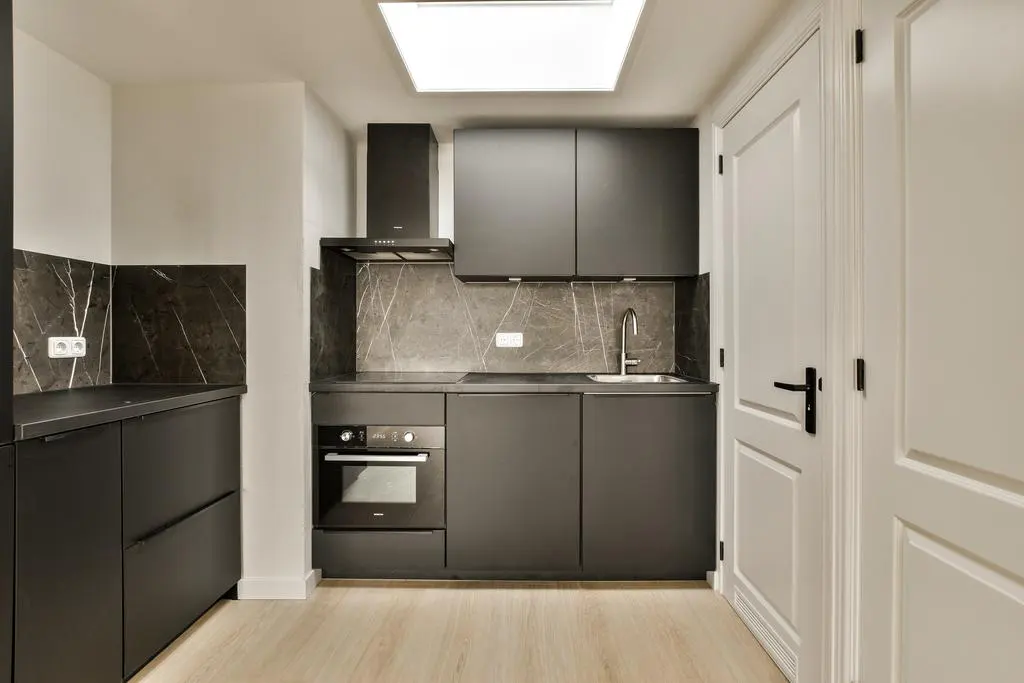 Modern kitchen with matte black cabinets, dark marble backsplash, built-in oven, and light wood flooring under a skylight.