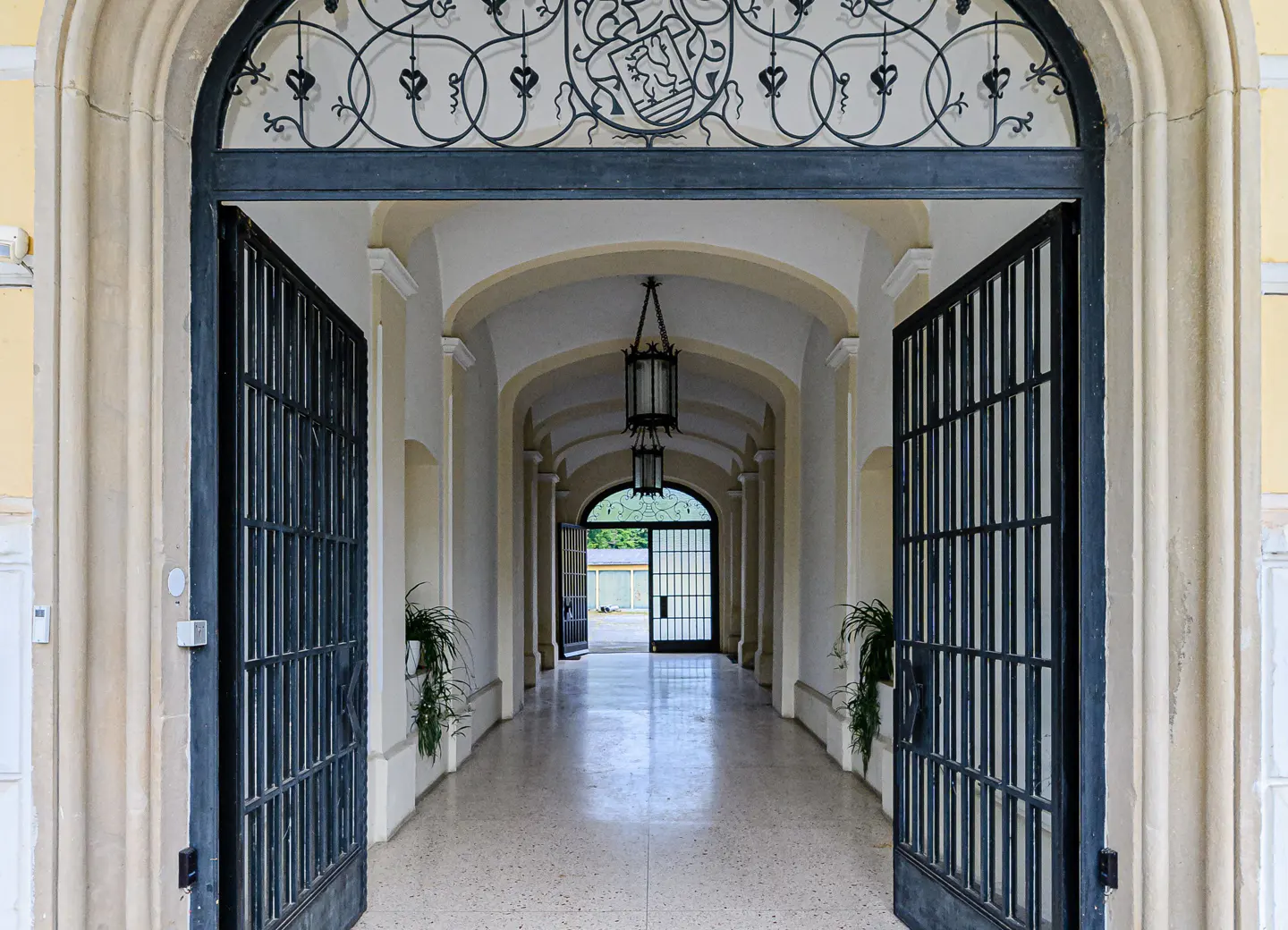 Arched entrance with open black iron gates leads to a long hallway with arched ceilings and hanging lanterns.