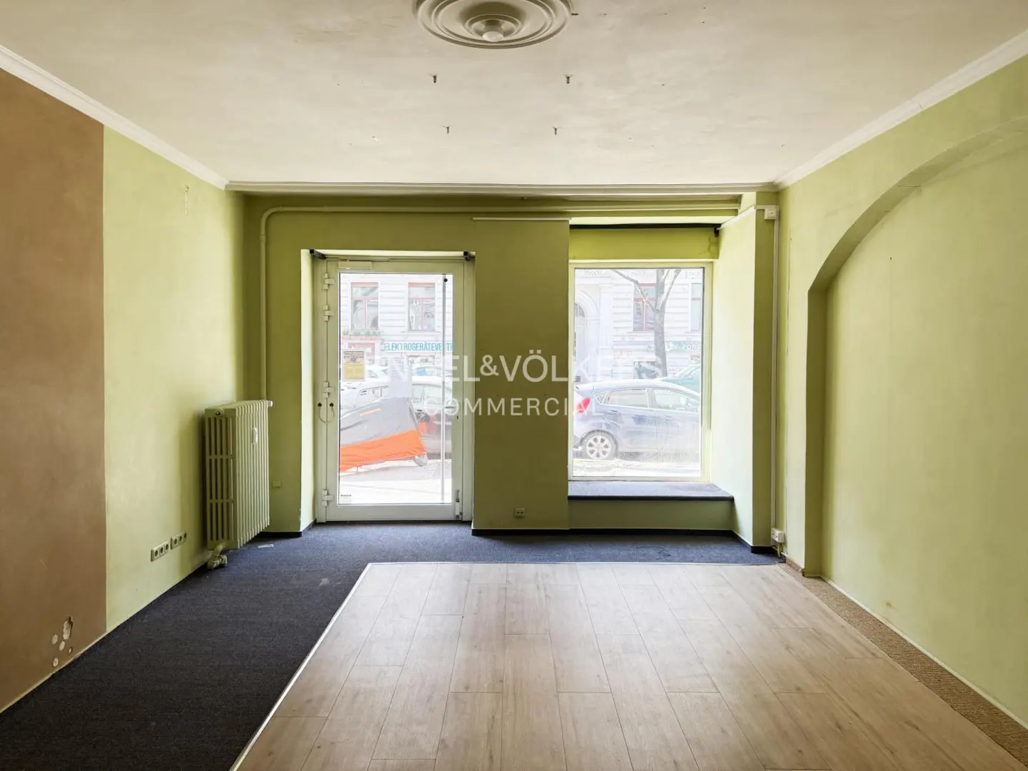 Empty commercial space with light green walls, wood floors, and dark carpet. A radiator sits by a window and glass door with the Engel & Völkers logo.