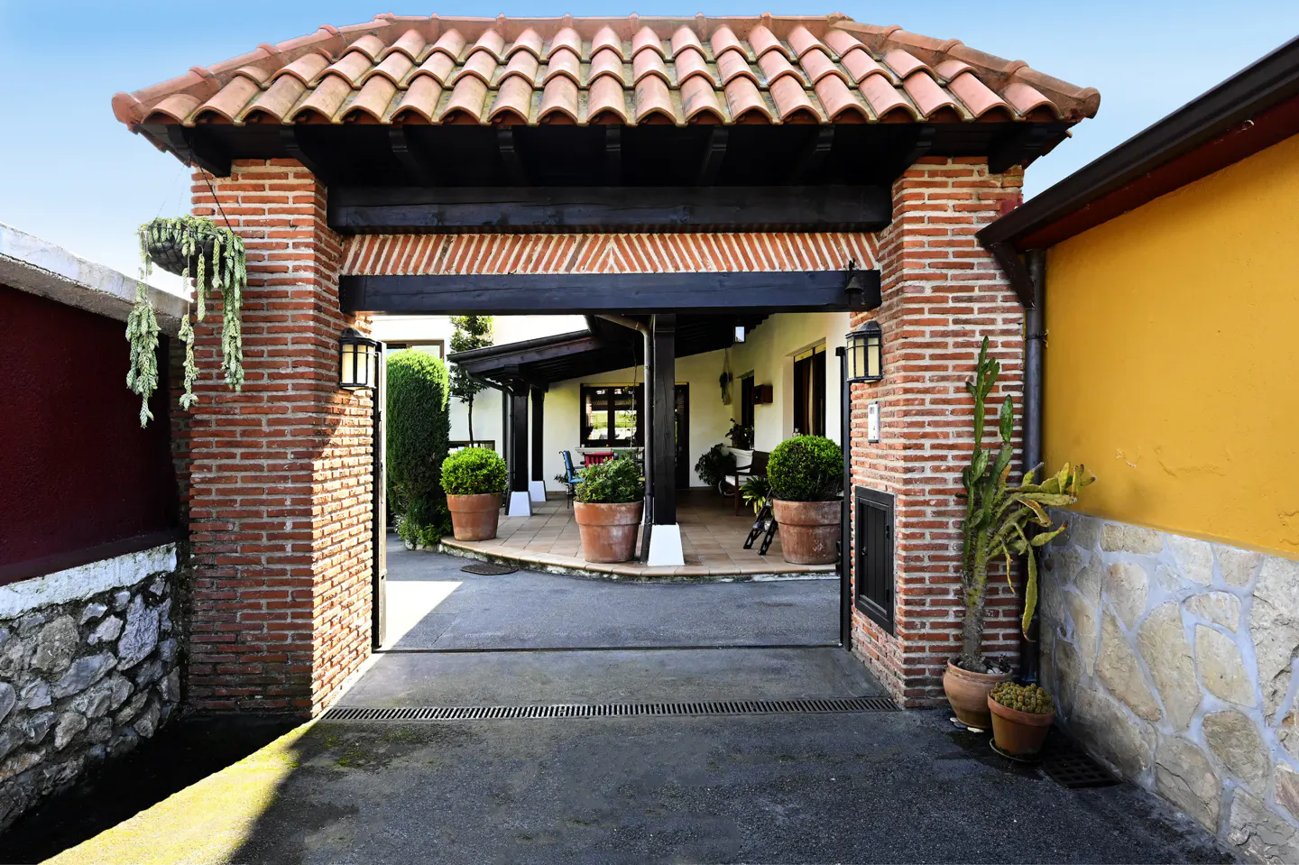 Brick archway with a red tile roof leads to a courtyard with potted plants and a white house with a covered porch.
