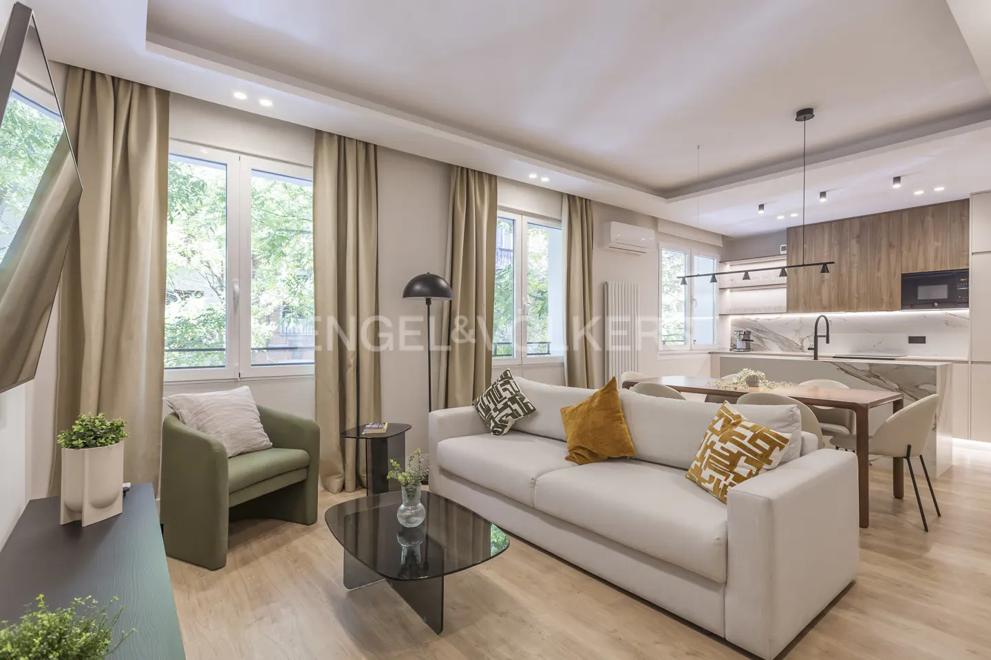 Bright, modern living room with a white sofa, green armchair, and open kitchen. Beige curtains frame the windows.