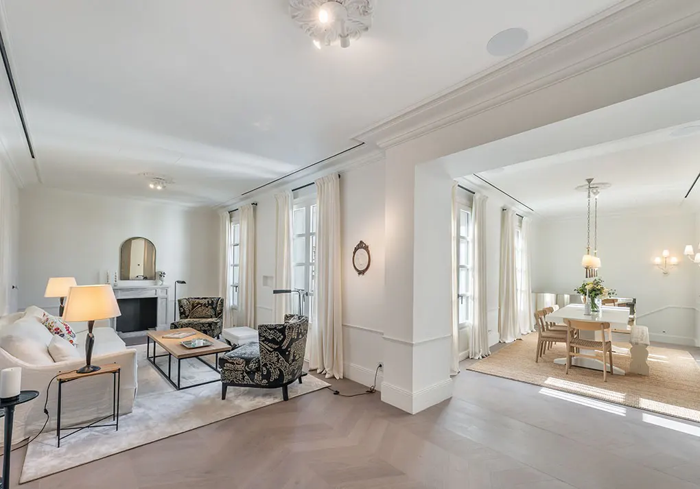 Bright, open-concept living and dining room with white walls, herringbone floors, and large windows with sheer curtains.