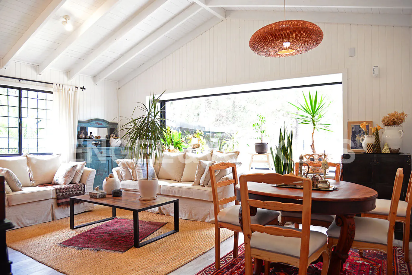 Bright living room with white walls, a brown woven light fixture, and a large window. A dining table and chairs are in the foreground.