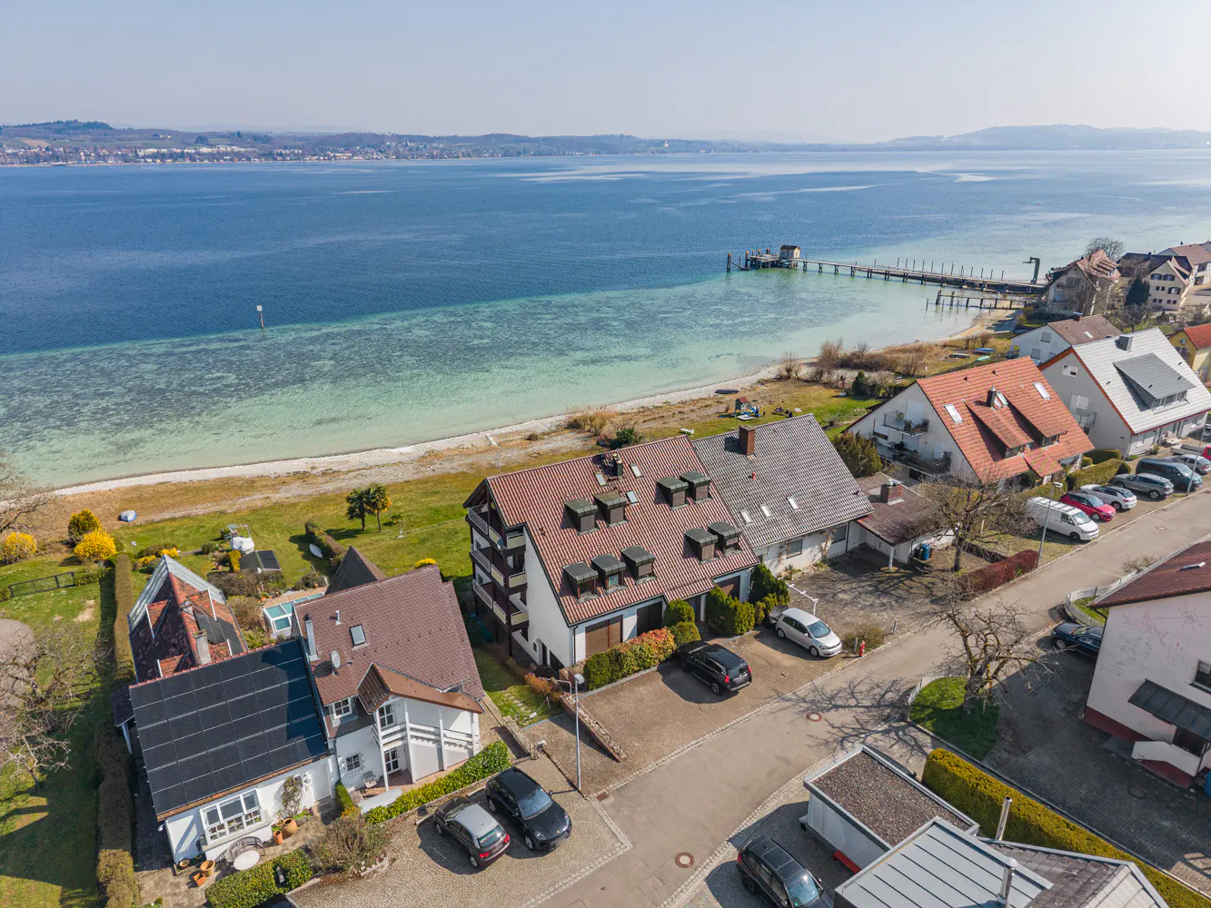 Aerial view of houses near a lake with a pier, cars parked in driveways, and green lawns.
