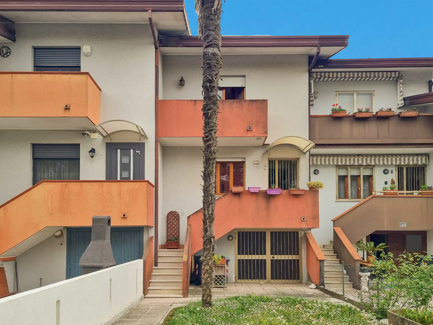 Three-story townhouses with orange balconies and staircases, set against a blue sky. A palm tree trunk is centered in the foreground.