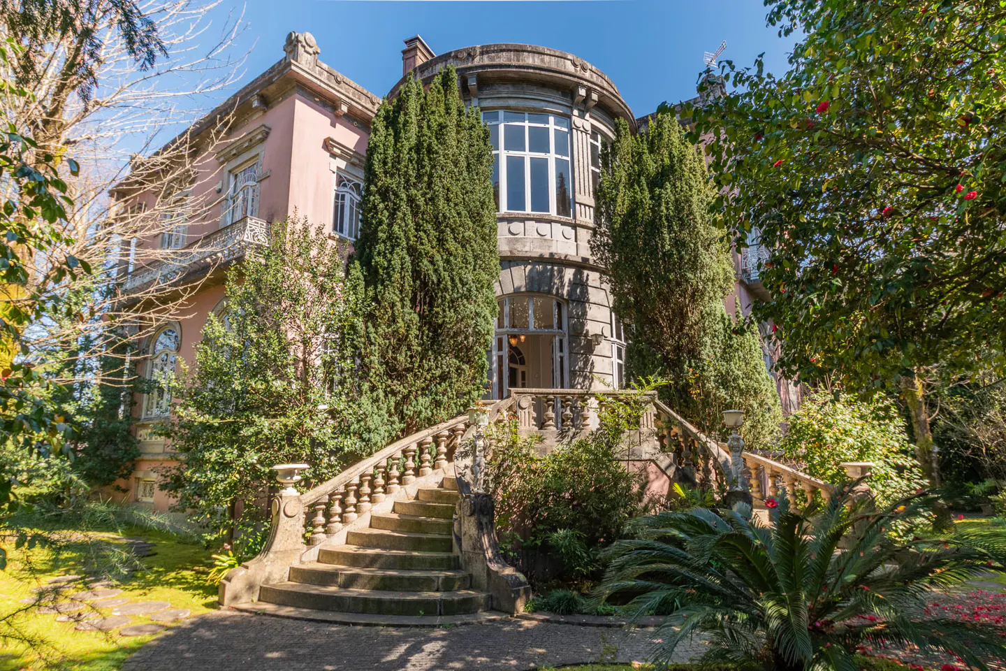 Exterior view of a pink mansion with a stone staircase leading to a rounded tower entrance, surrounded by lush greenery.