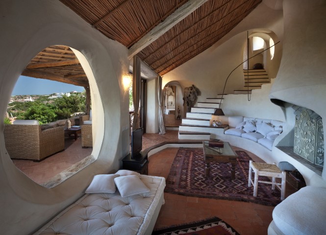 Interior view of a unique home with white walls, a thatched roof, and a large, round window overlooking a patio and landscape. A staircase leads to an upper level. [AI-generated text]