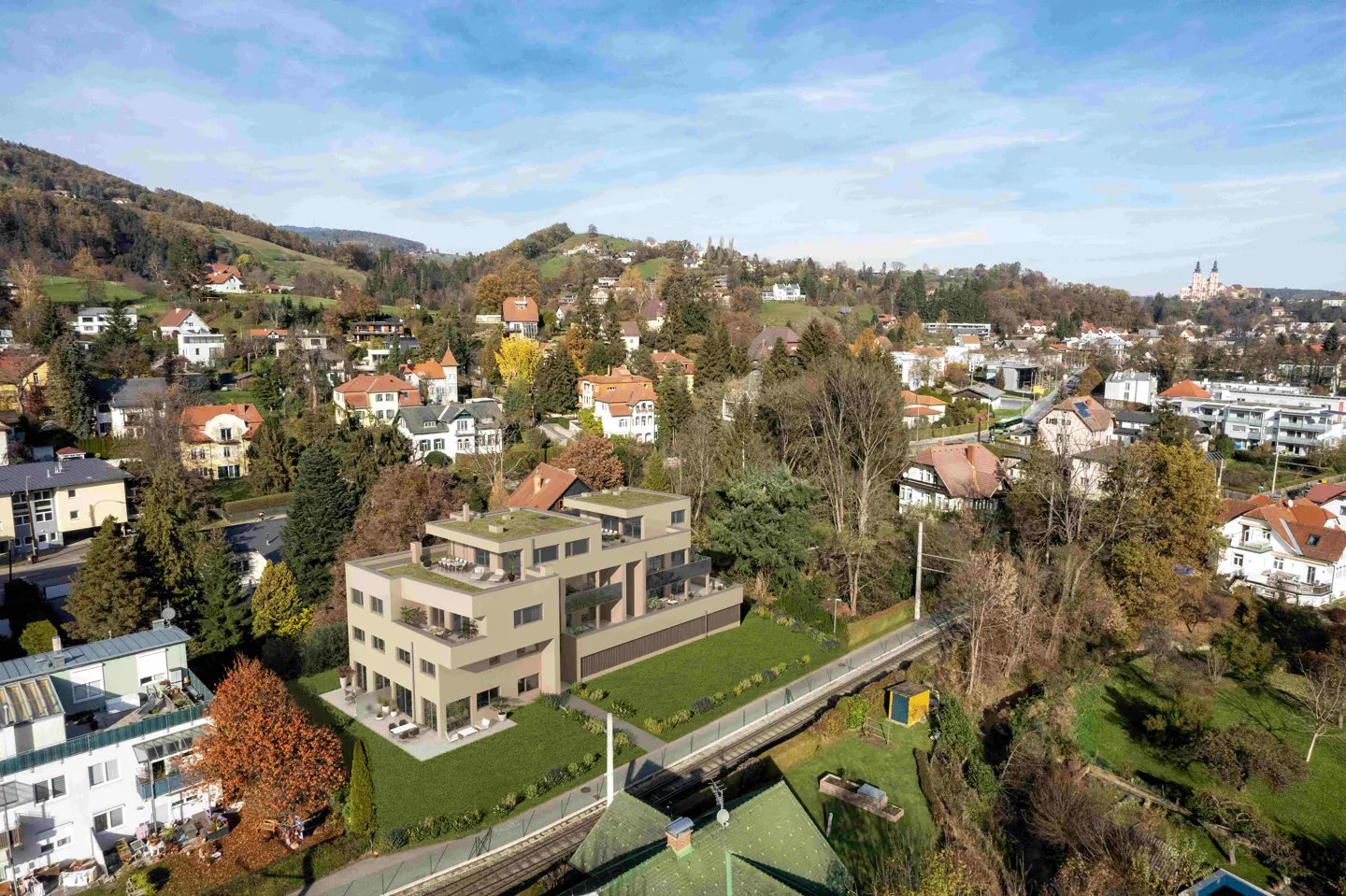 Aerial view of a modern, beige apartment building with green roofs, set in a lush, hilly landscape with scattered houses and a blue sky.