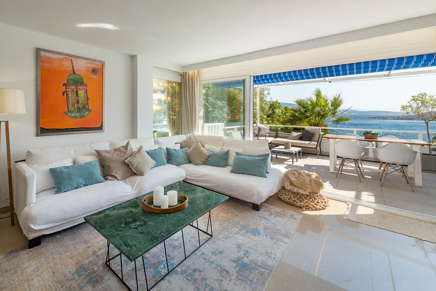 Bright living room with white sofas, teal pillows, green marble coffee table, and a balcony overlooking the blue sea.