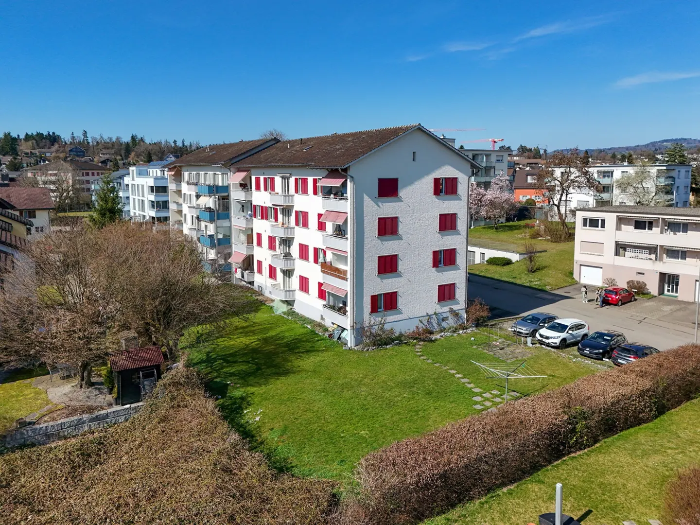 Exterior view of a white apartment building with red shutters and awnings, surrounded by green lawn and trees. Cars are parked nearby.