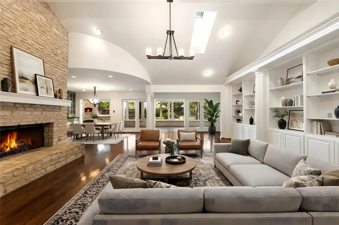 Bright, open living room with stone fireplace, hardwood floors, and white built-in bookshelves. A gray sectional sofa faces the fireplace.
