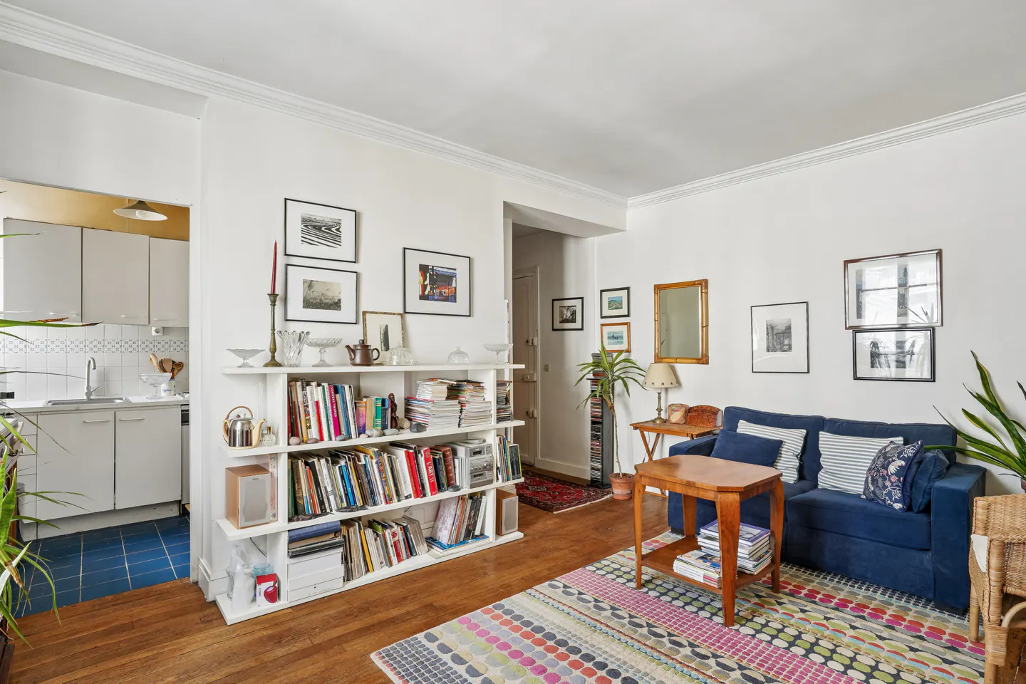 Bright living room with white walls, wood floors, and a blue sofa. A bookshelf is filled with books and decor. Kitchen visible in the background.