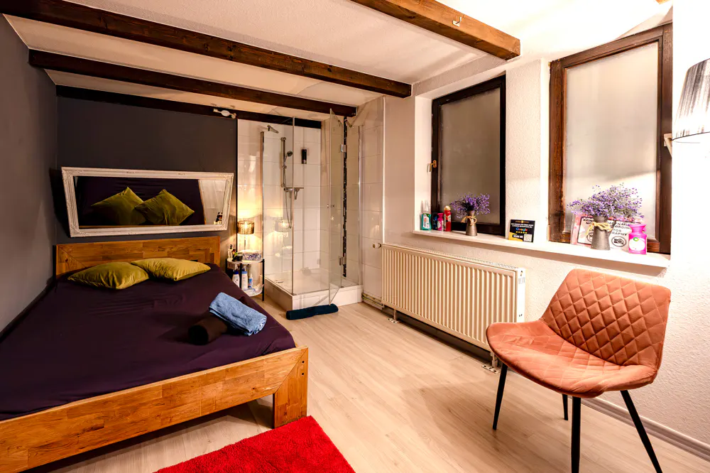 Cozy bedroom with wood beams, a wooden bed frame, purple sheets, and a glass shower. A brown chair sits near a window.