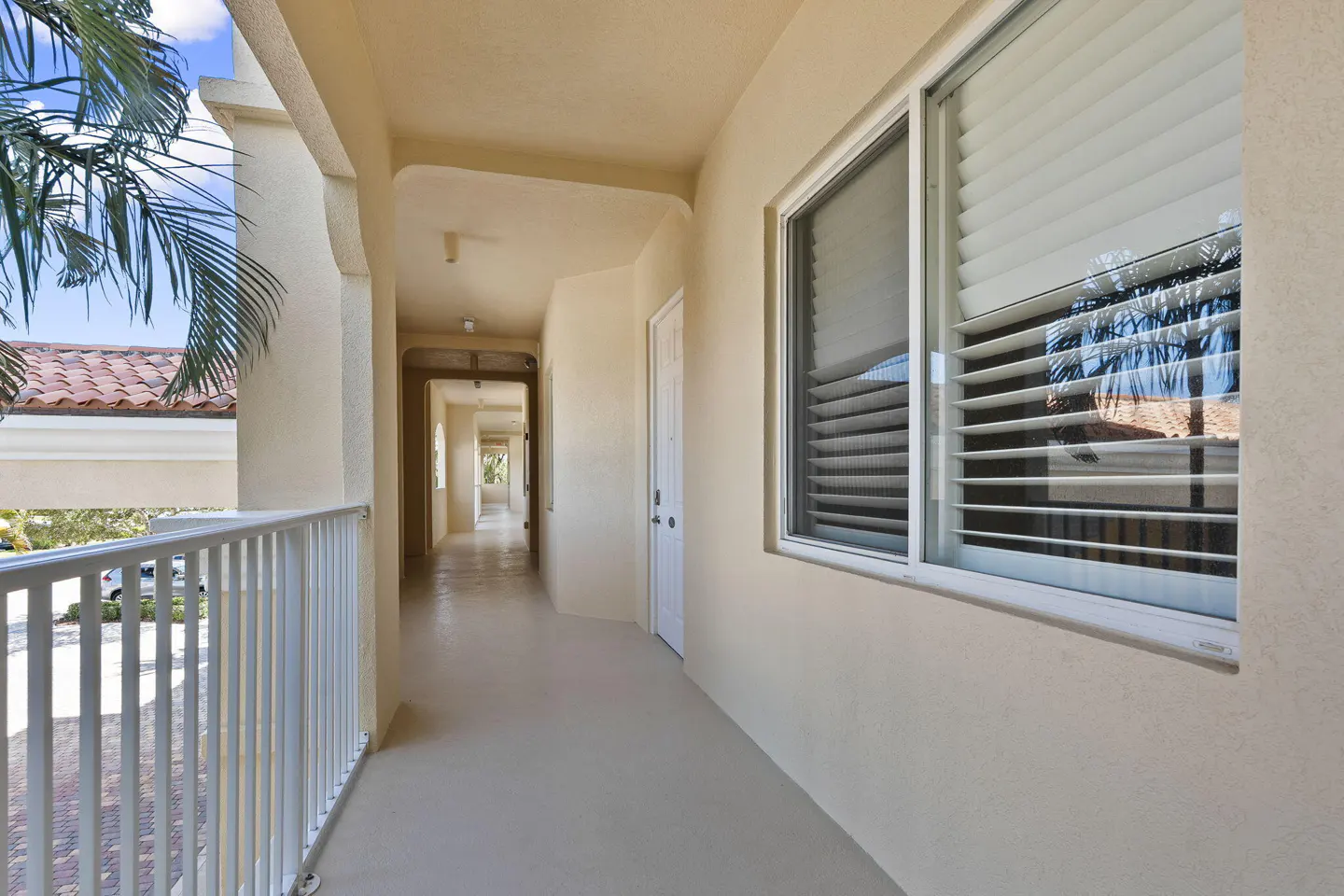 A covered outdoor walkway with cream walls, a white railing, and a window with white blinds overlooking a tiled roof.