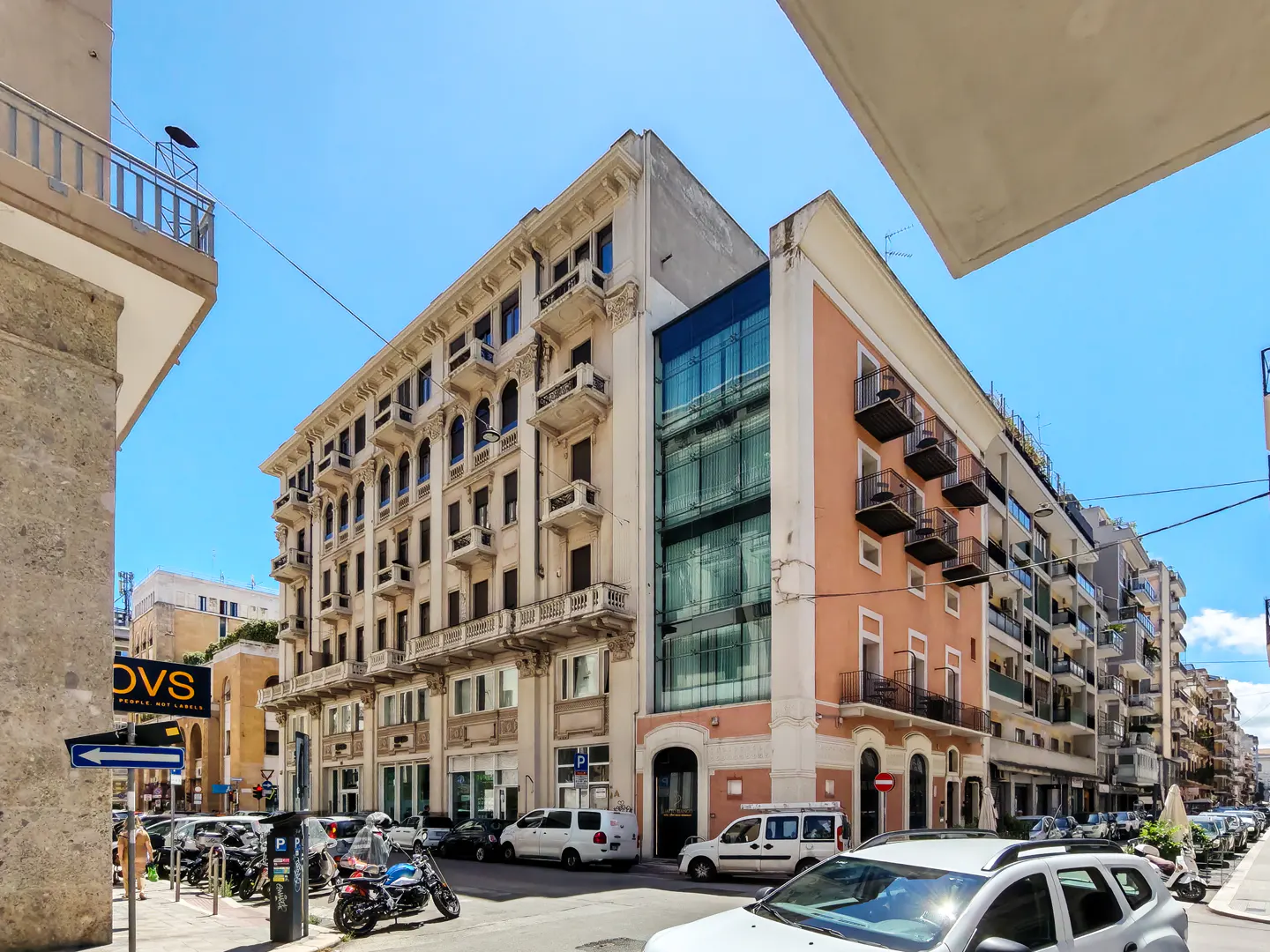 A street view of a historic beige building next to a modern glass-fronted structure under a clear blue sky.