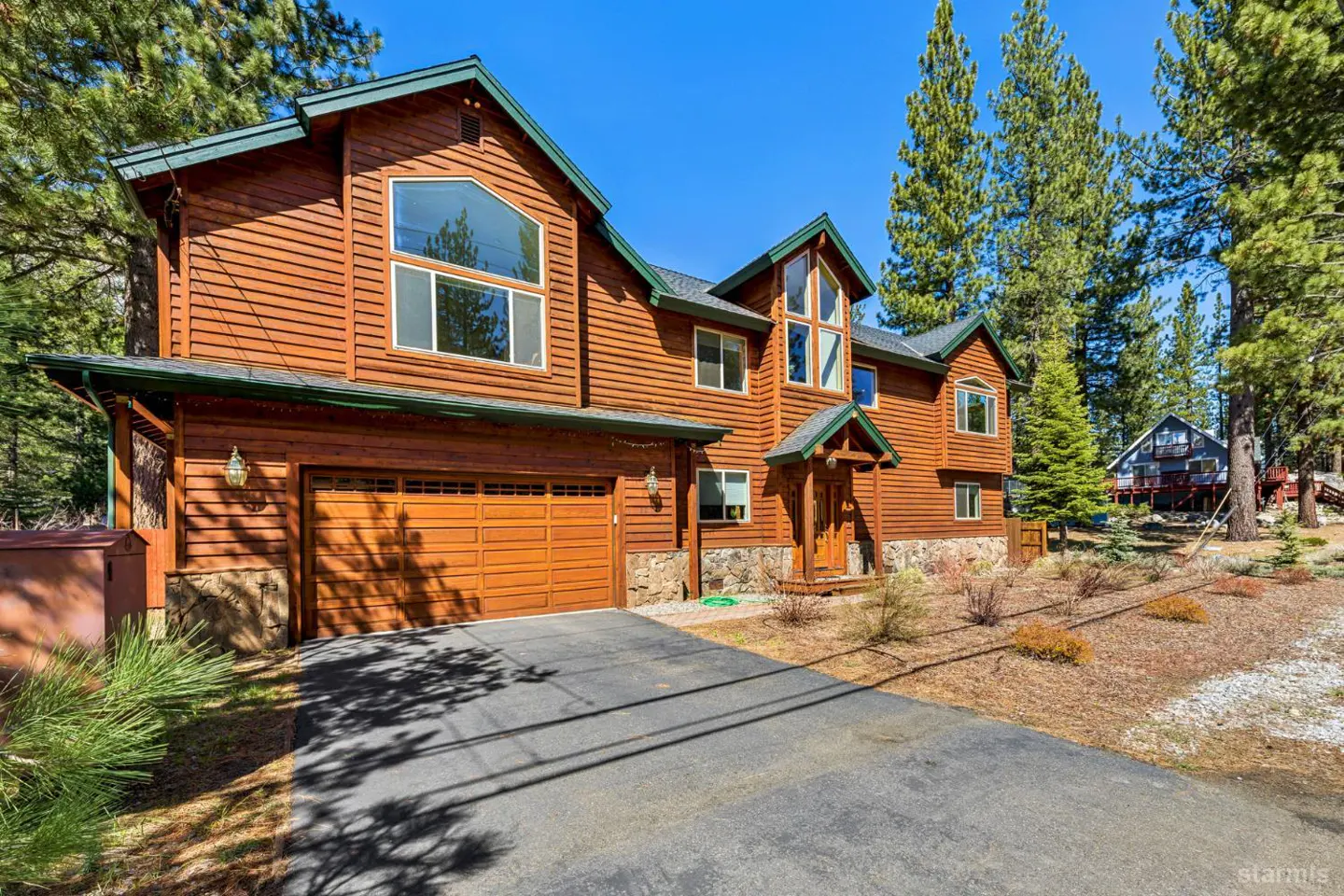 A two-story wood cabin with a garage and a driveway surrounded by tall trees.