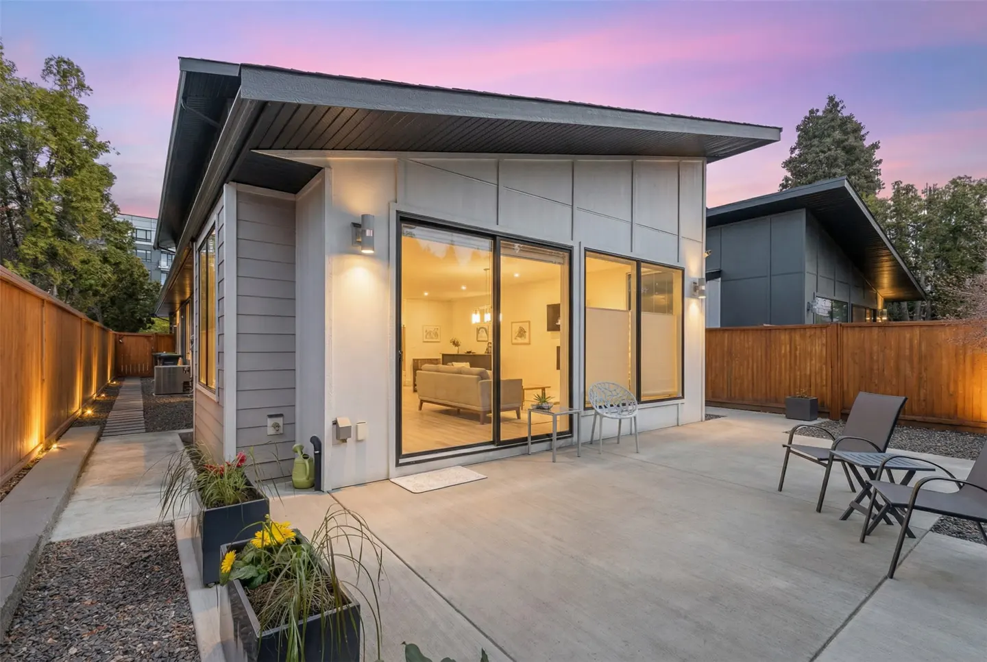 Modern home exterior with a concrete patio, chairs, and sliding glass doors revealing a living room at dusk.