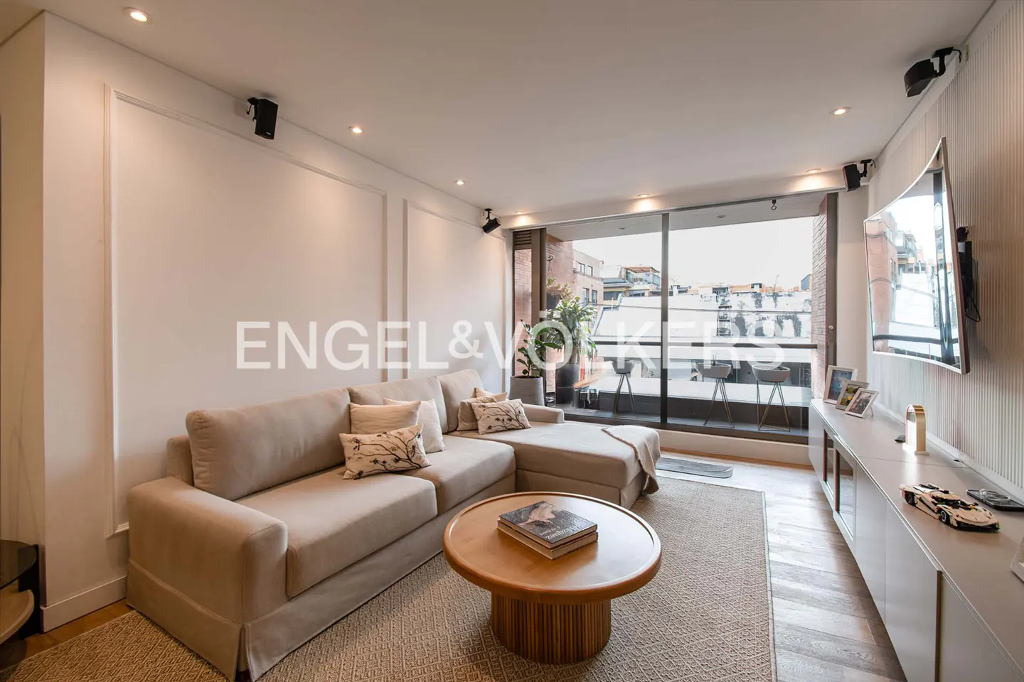 Living room with beige sectional sofa, round wood table, and balcony with city view. TV and console table on the right.