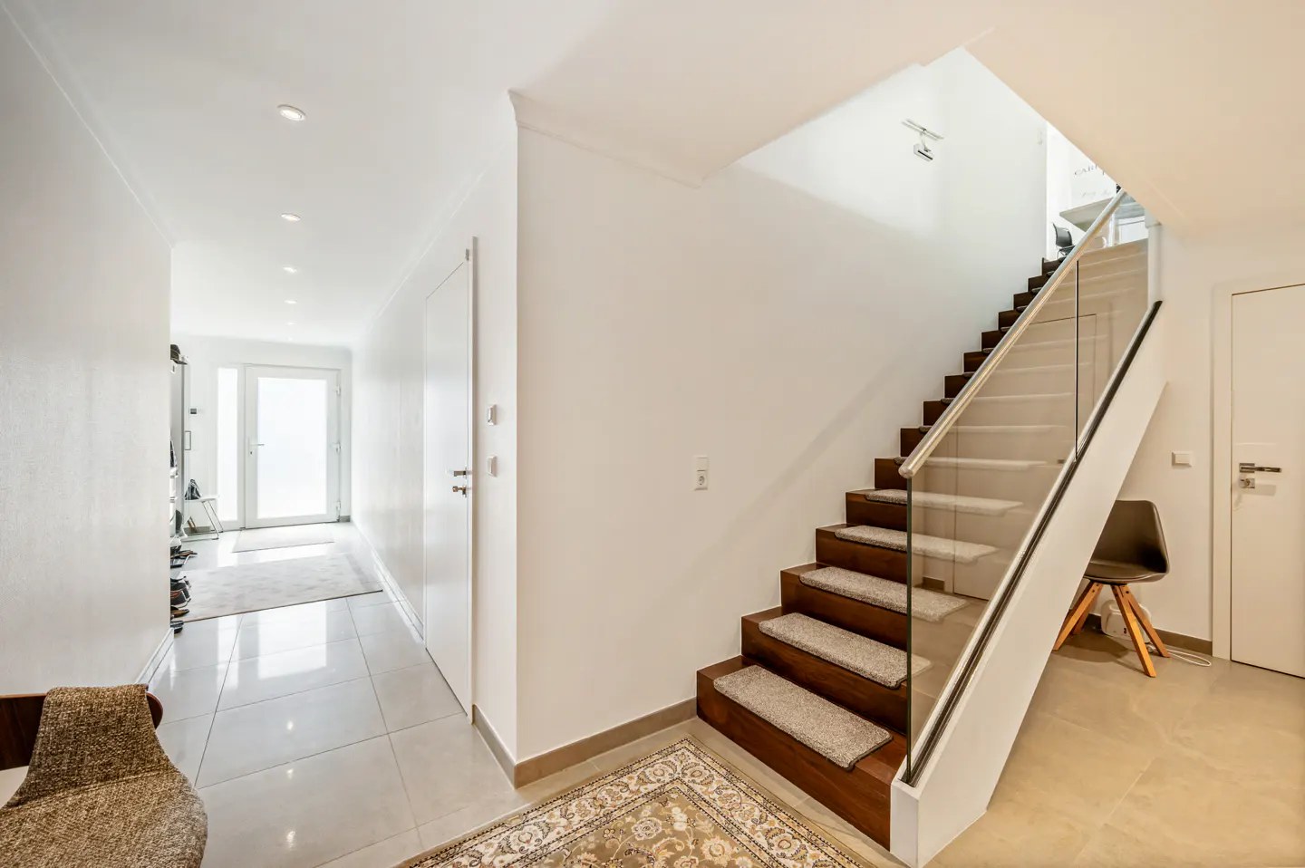 Bright hallway with white walls, tiled floor, and a wooden staircase featuring carpet treads and a glass railing.