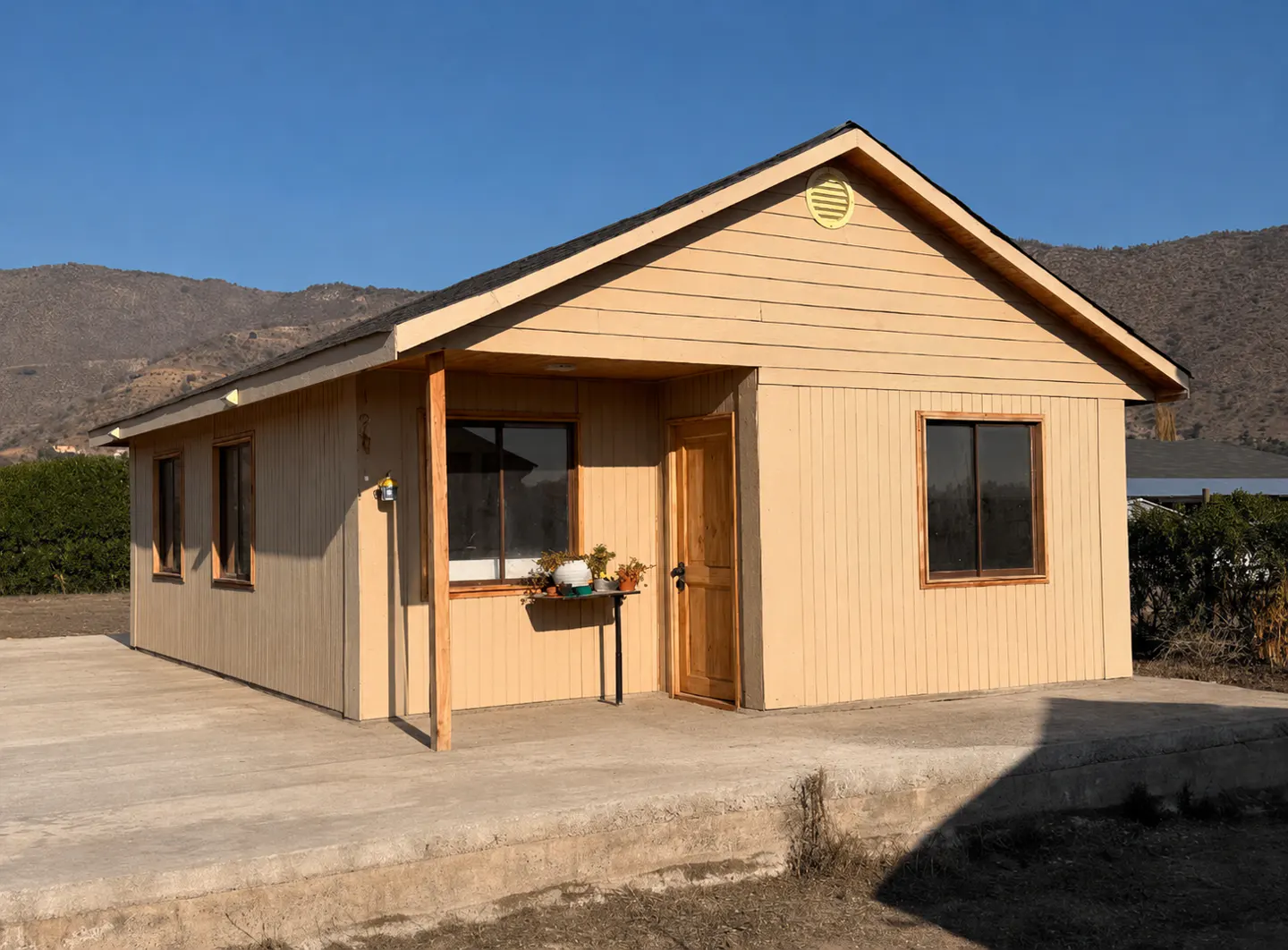 A single-story tan house with a concrete porch, wooden door, and dark-framed windows, set against a backdrop of mountains.