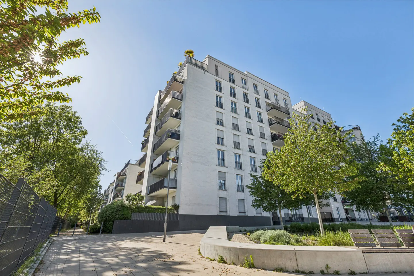 Modern white apartment building with black balconies, set against a blue sky and green trees.
