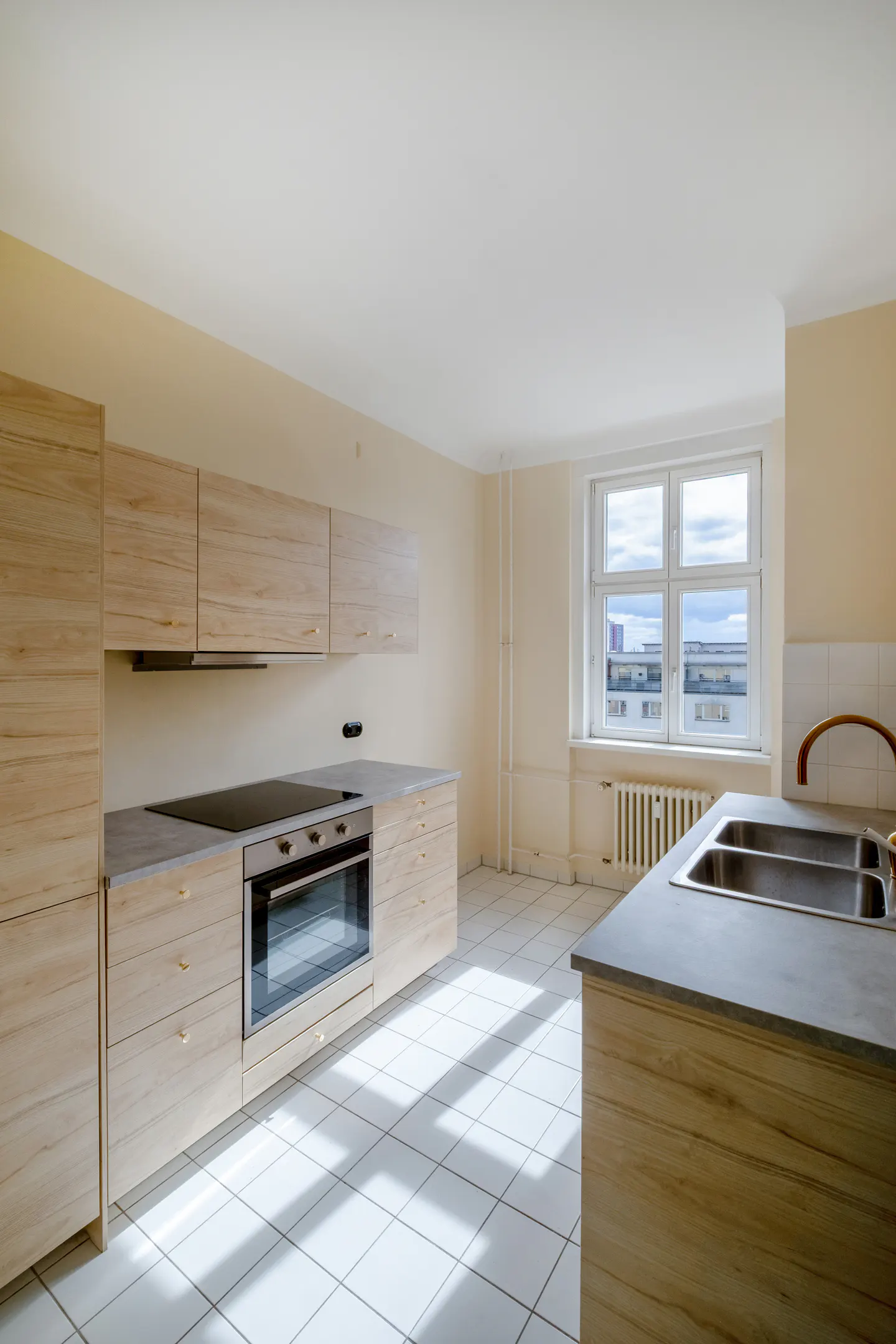 Bright kitchen with light wood cabinets, gray countertops, and white tile floor. A window overlooks a cityscape.