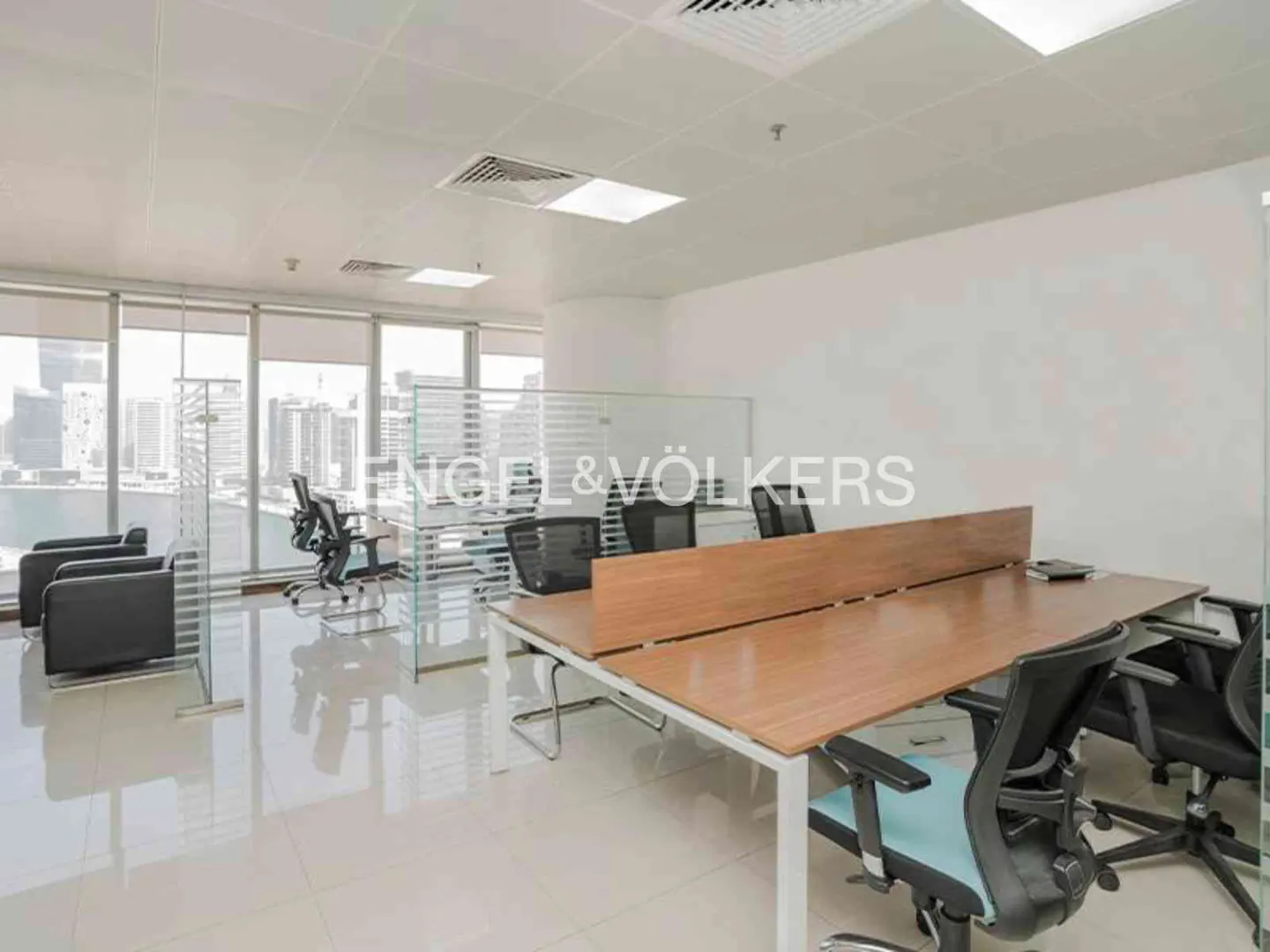 Bright office space with desks, chairs, and glass partitions. Large windows offer a city view. The floor is shiny and reflective.
