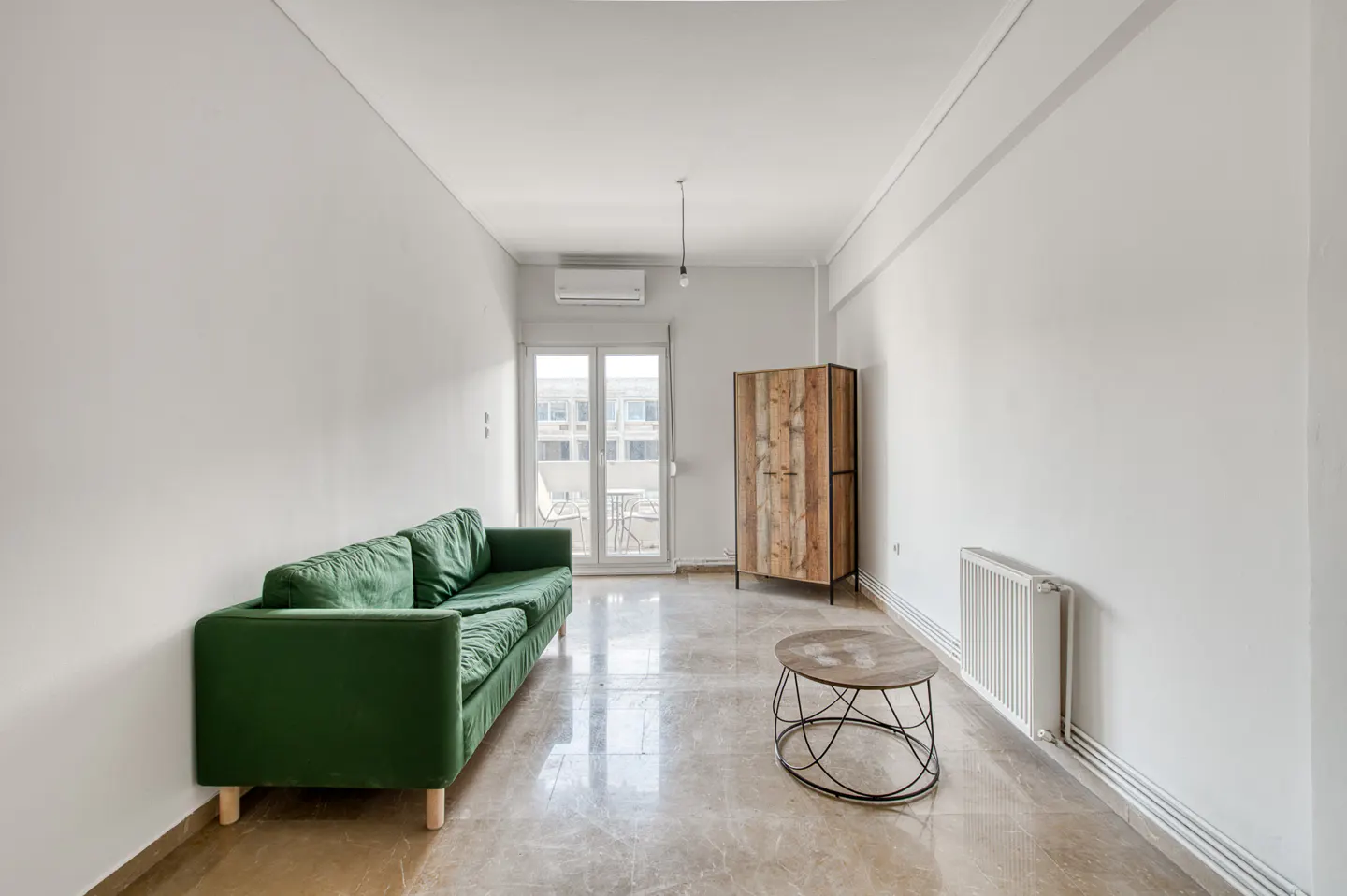 Bright living room with a green sofa, wooden cabinet, and a small table on a marble floor. Balcony doors let in natural light.