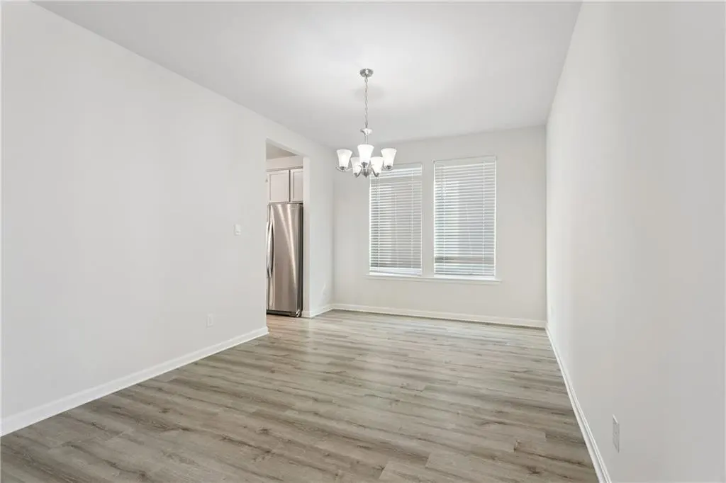 Bright, empty dining room with gray wood floors, white walls, chandelier, and view into kitchen with stainless steel refrigerator.