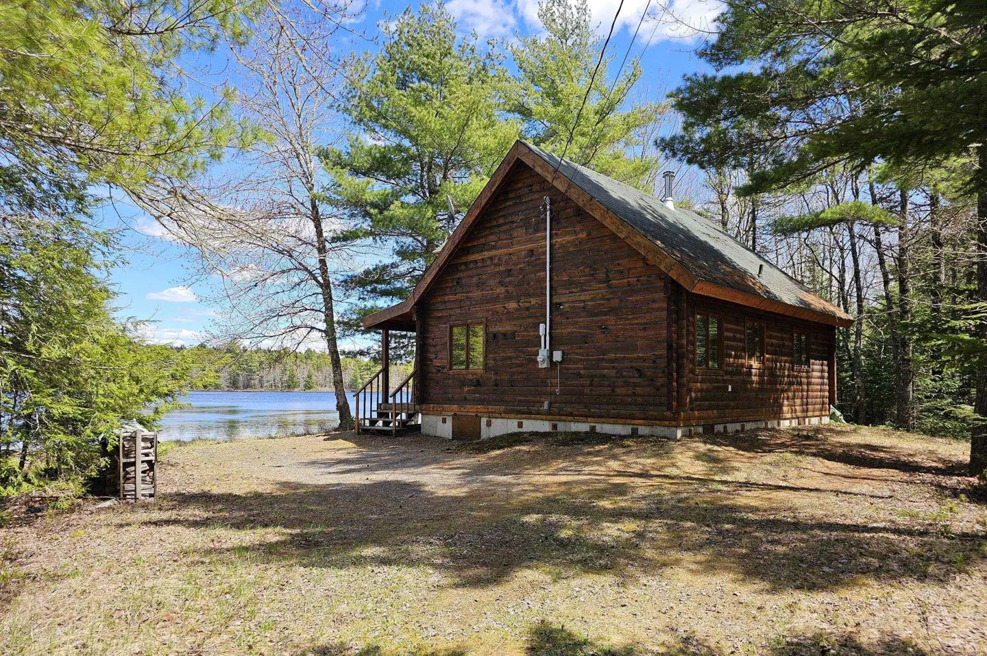 A rustic brown log cabin with a green roof sits near a lake under a blue sky with scattered clouds. Tall green trees surround the cabin.