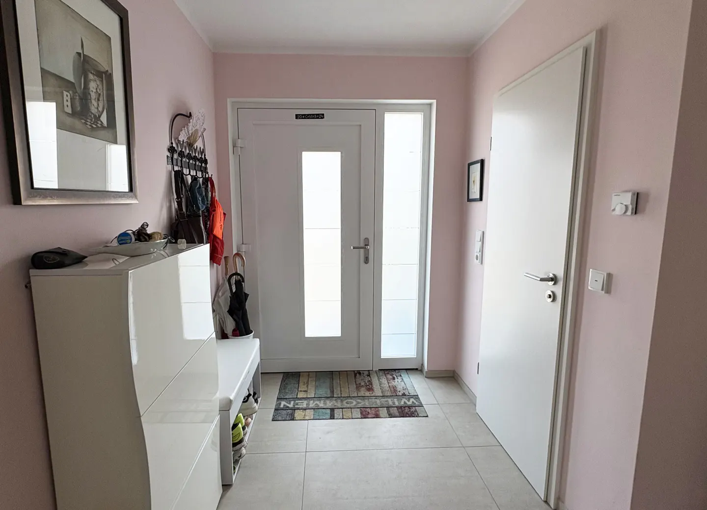 Hallway with pink walls, white doors, and light tile flooring. A white cabinet and bench sit to the left of the front door.