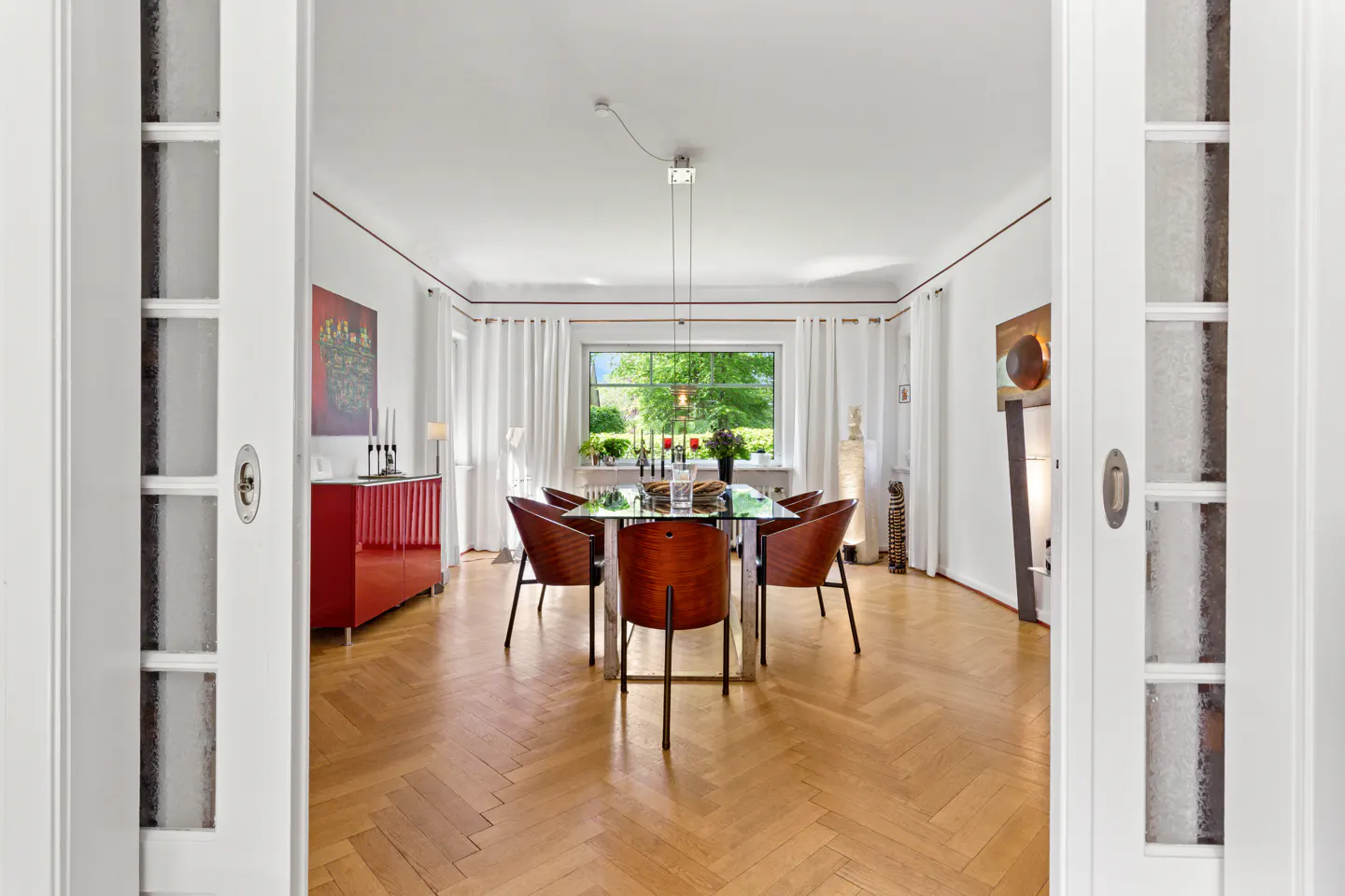 View through white doors into a dining room with a glass table, wood chairs, and herringbone wood floors. A red cabinet sits against the wall.