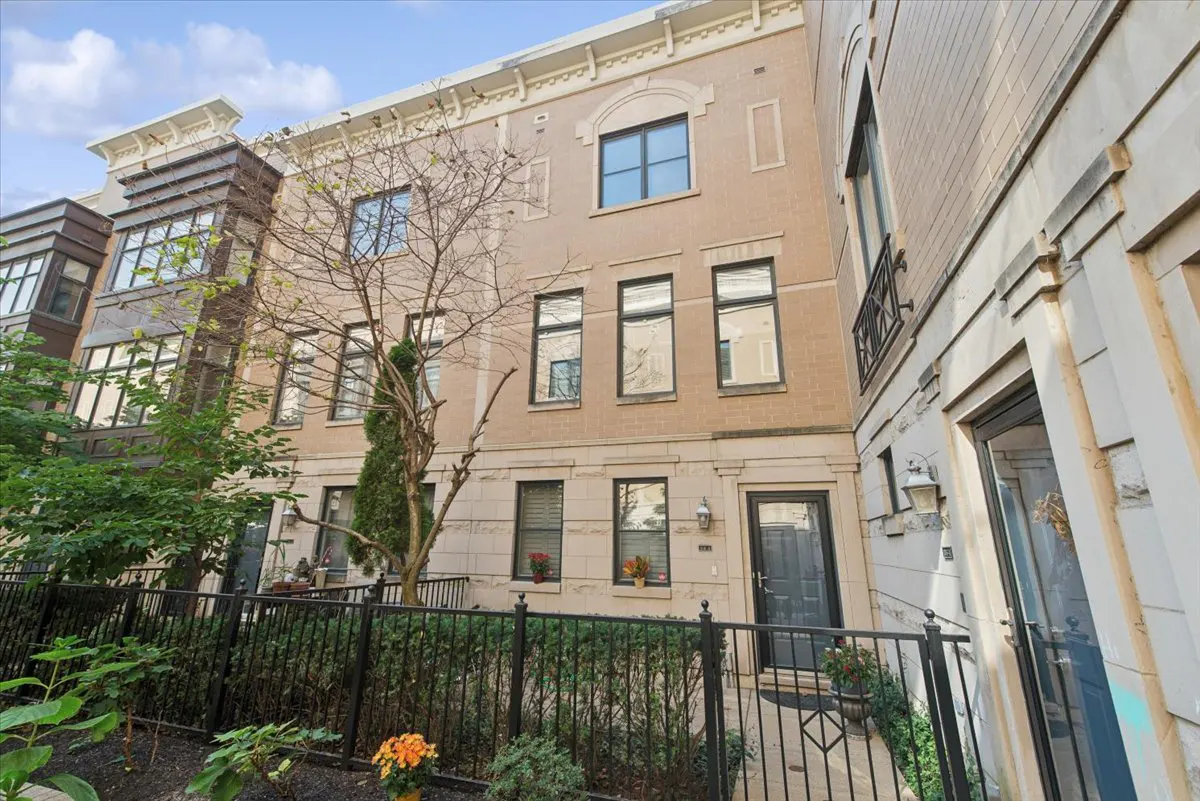 Three-story tan brick townhouse with black trim and a small front yard with a black metal fence.