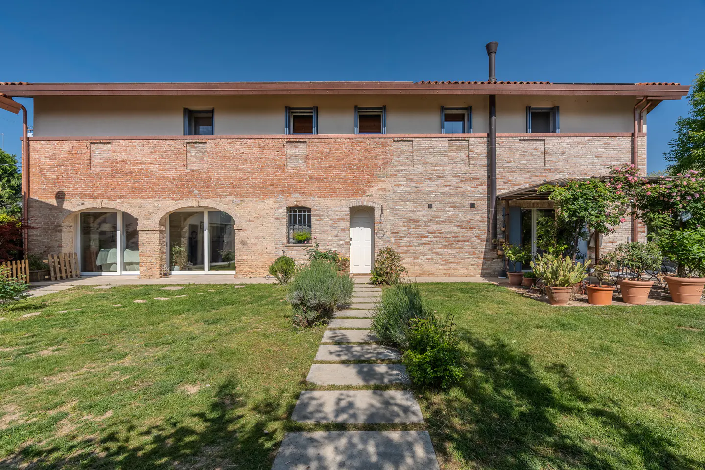 Two-story brick house with a green lawn and stone pathway leading to the front door. Potted plants and flowers add color to the yard.