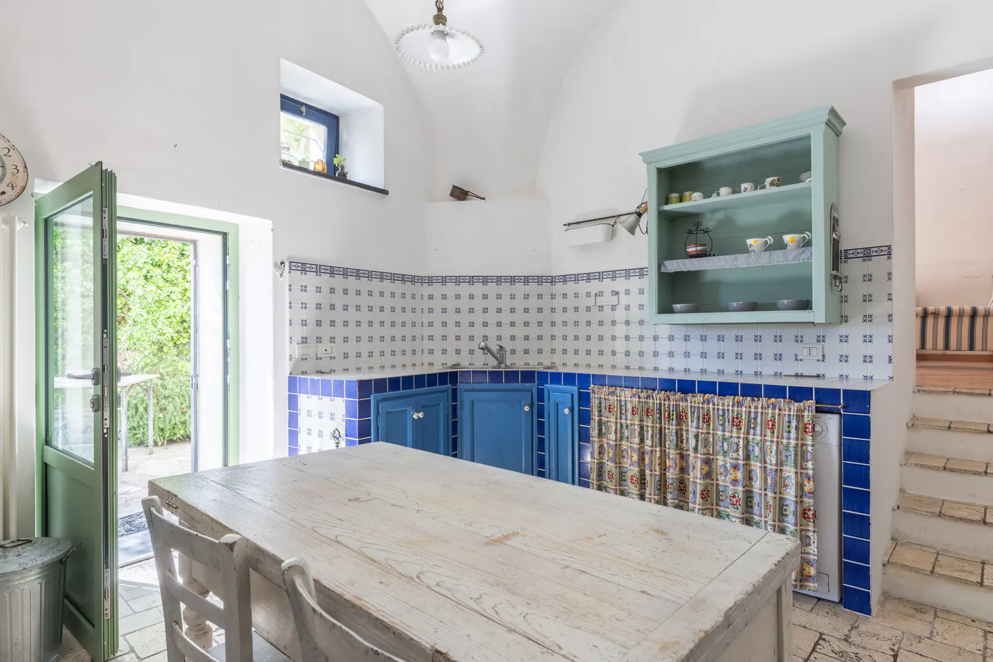 A bright kitchen with white walls, blue cabinets, and a large wooden table. An open green door leads to a garden.