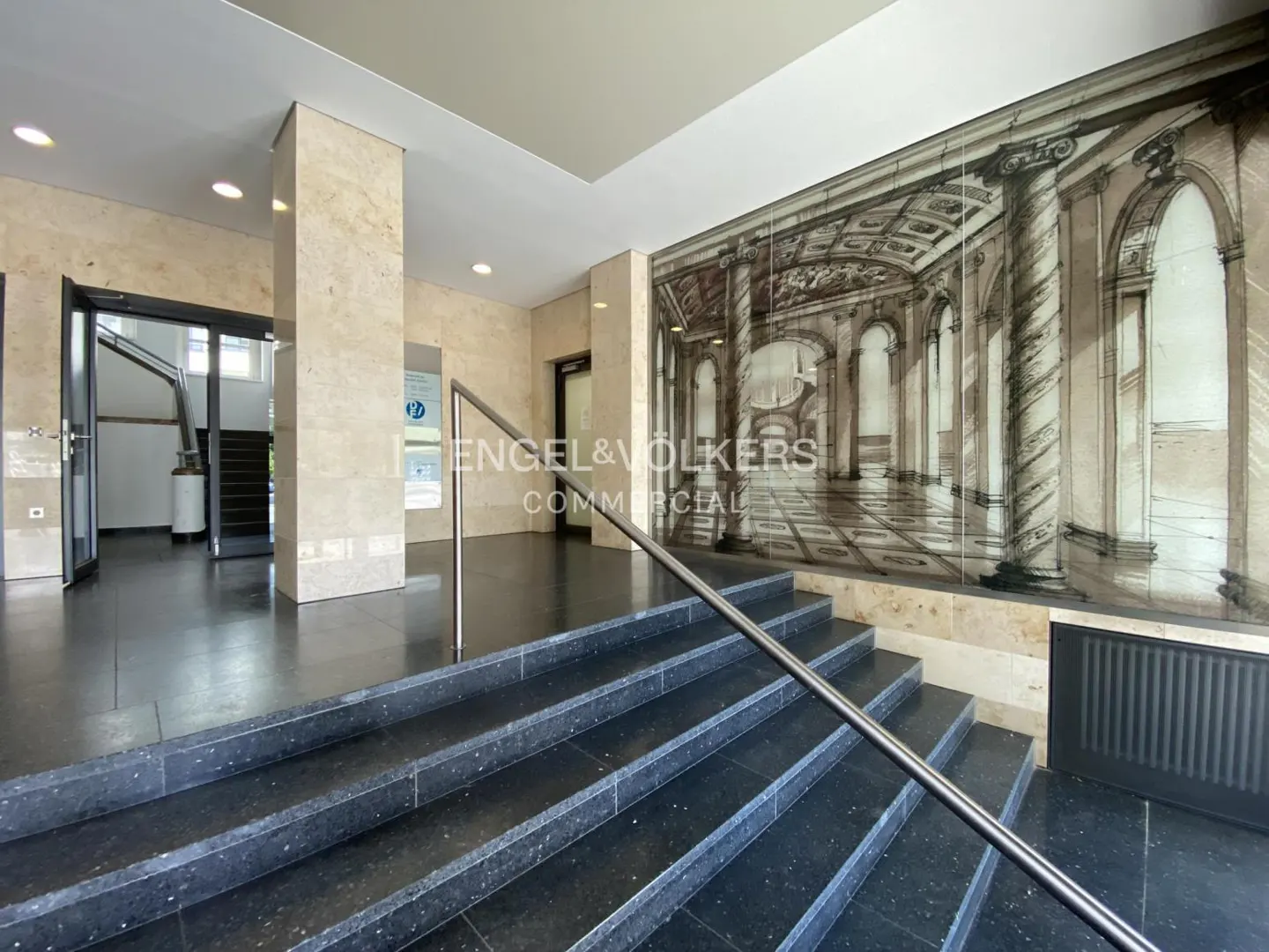 Office lobby with black stairs, metal railing, and marble walls. An Engel & Völkers Commercial sign is visible. A mural of a building interior is on the wall.