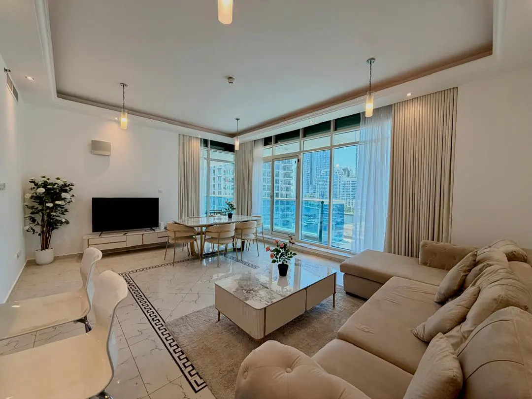 Modern living room with beige sectional sofa, marble coffee table, dining area, and floor-to-ceiling windows with city view.