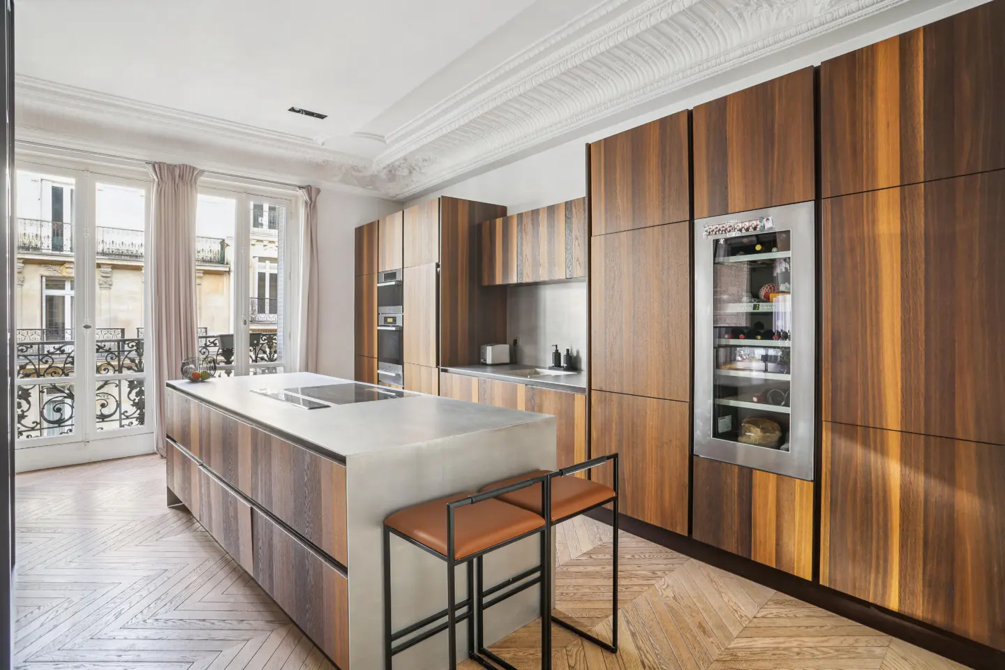 A modern kitchen with wood cabinets, a concrete island with two brown leather stools, and a wine fridge. A large window overlooks a city street.