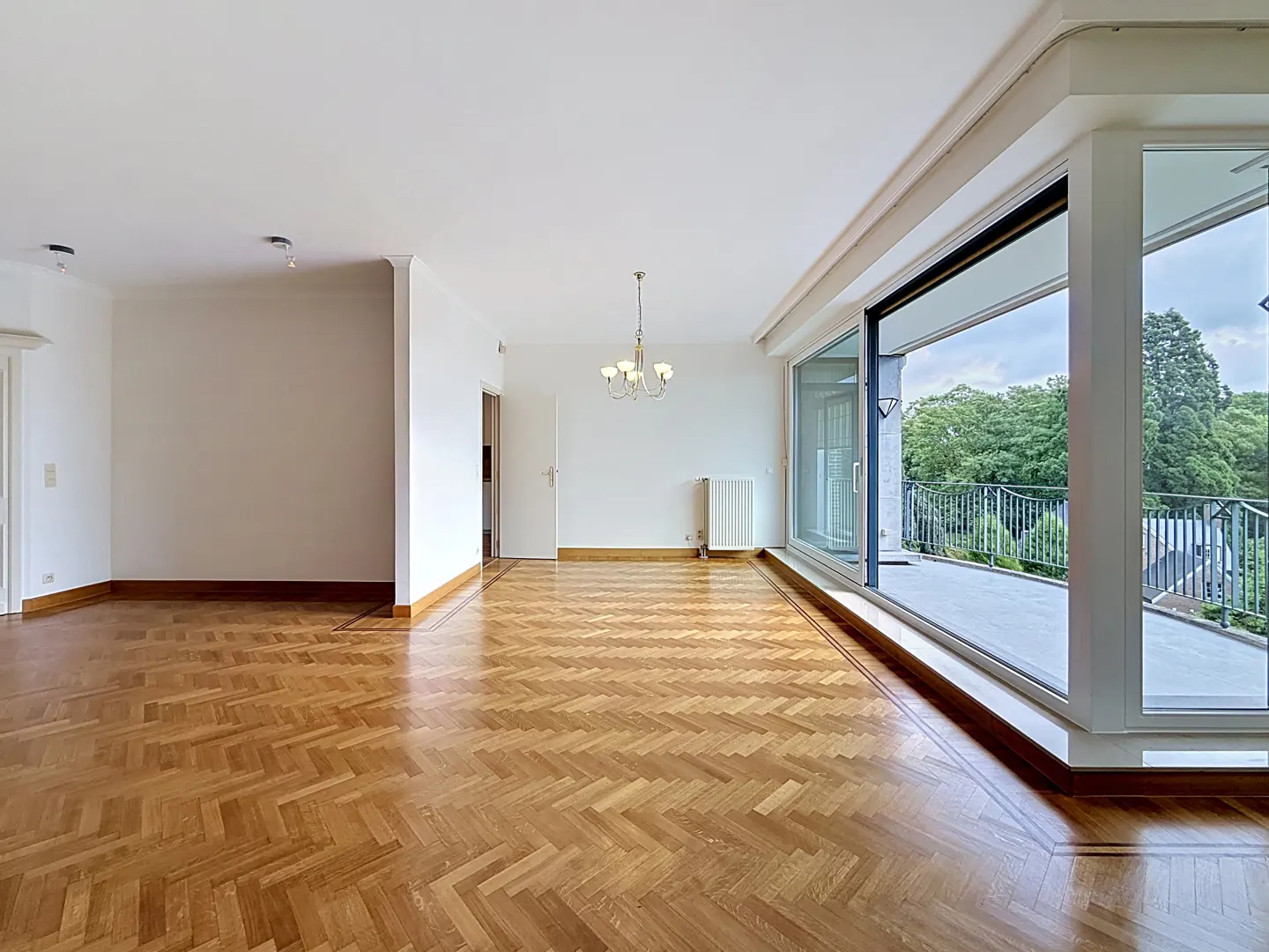 Bright, empty living room with herringbone wood floors, a classic chandelier, and large sliding doors opening to a balcony.