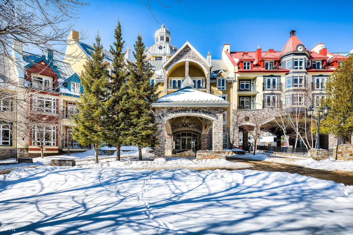 Colorful buildings with snow-covered roofs and trees on a sunny day. "Le Sommet des Neiges" is written above the entrance.