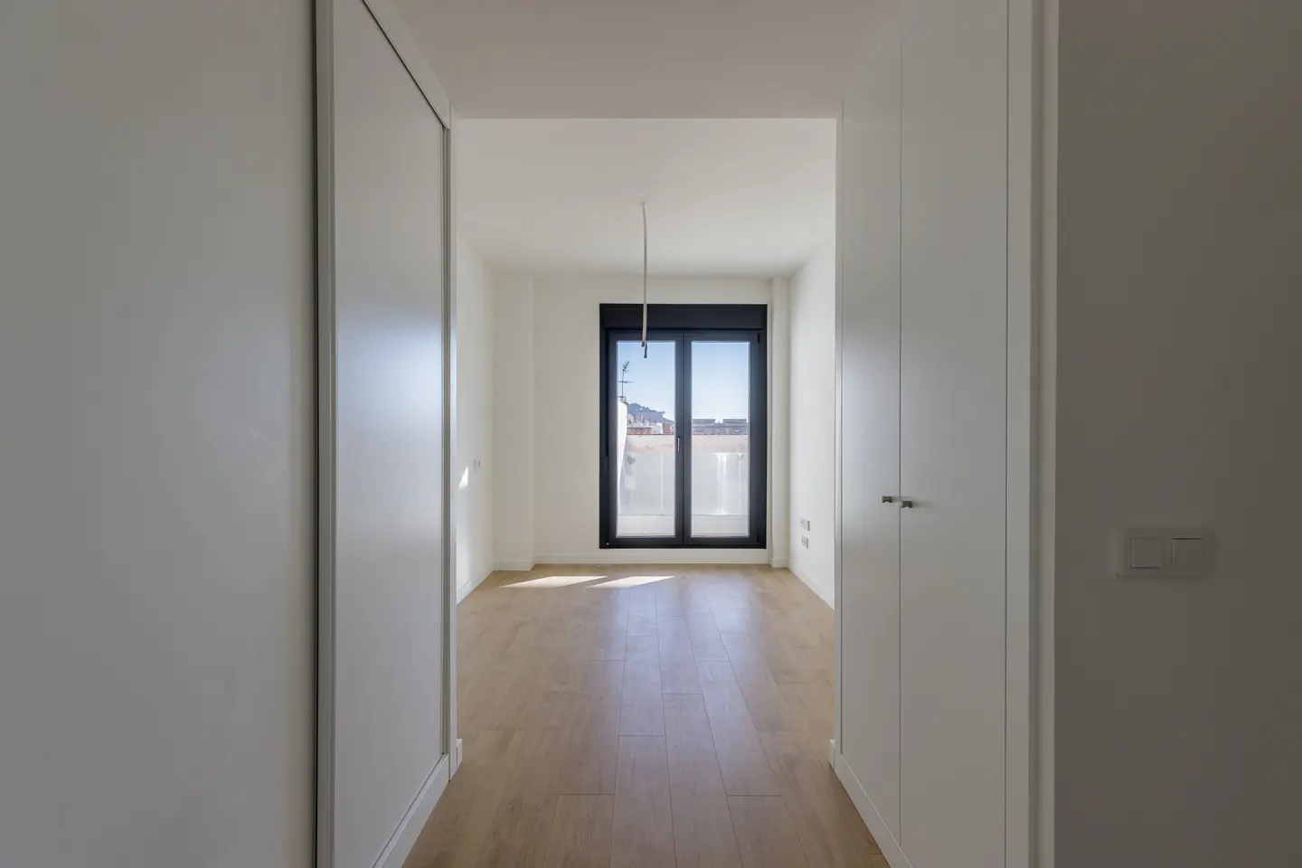 A bright, empty room with light wood floors and white walls, viewed through a doorway framed by white closets. A window with a black frame is at the far end.
