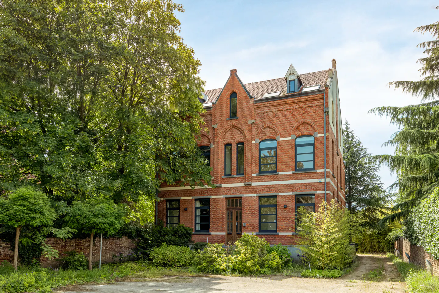 A three-story red brick house with dark window frames, surrounded by lush green trees and a gravel driveway.