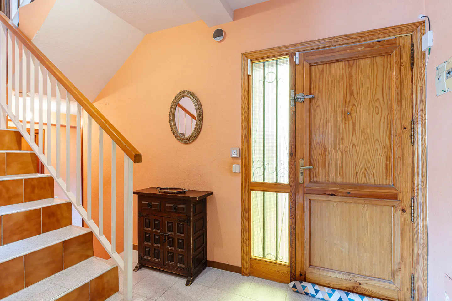 Entryway with wooden door, stairs, and cabinet. Peach walls, white trim, and a round mirror add warmth.