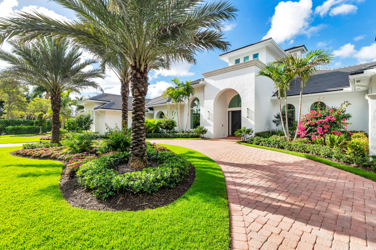 A white, two-story house with a brick driveway and palm trees. The sky is blue with some clouds.