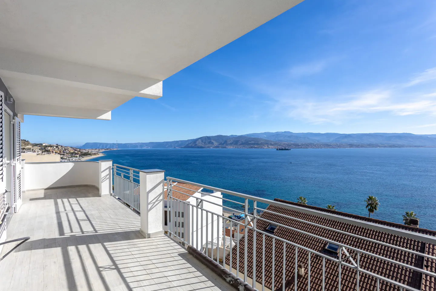Balcony view of blue ocean and sky. White railings cast shadows on the tiled floor. Distant mountains and coastline visible.