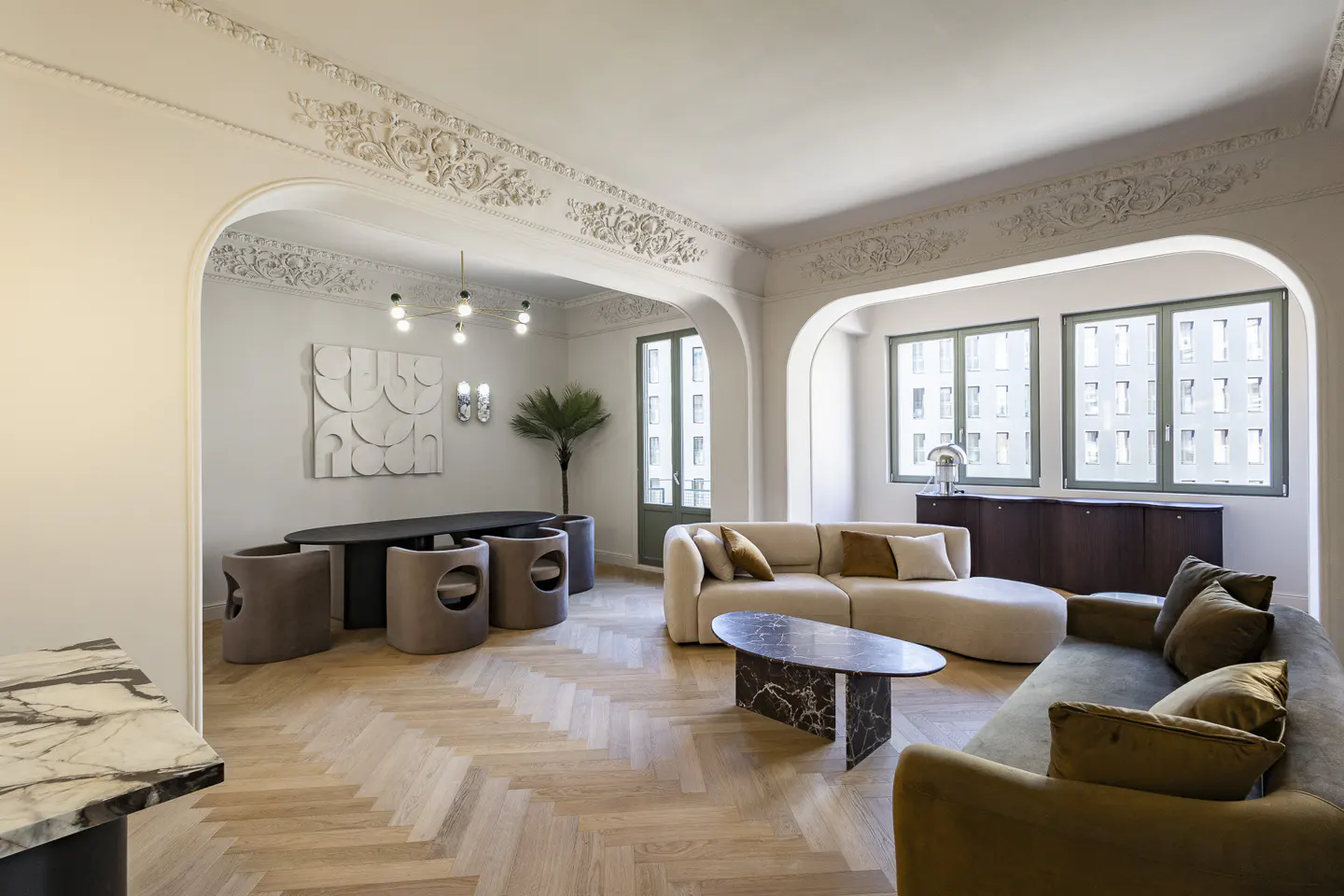 Bright, modern living room with herringbone wood floors, arched doorways, and ornate ceiling trim. Sofas, table, and chairs furnish the space.