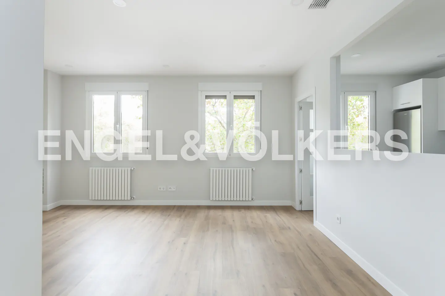 Bright, empty room with light wood floors, white walls, two windows, and radiators. Kitchen visible through opening. Engel & Volkers logo.
