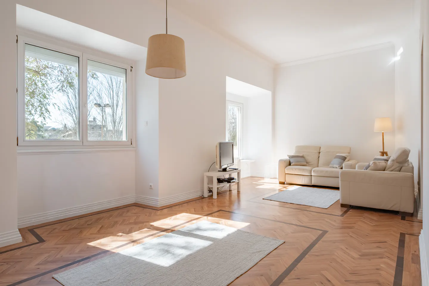 Bright, airy living room with hardwood floors, white walls, and two beige sofas. Sunlight streams through the windows. A beige pendant lamp hangs from the ceiling.