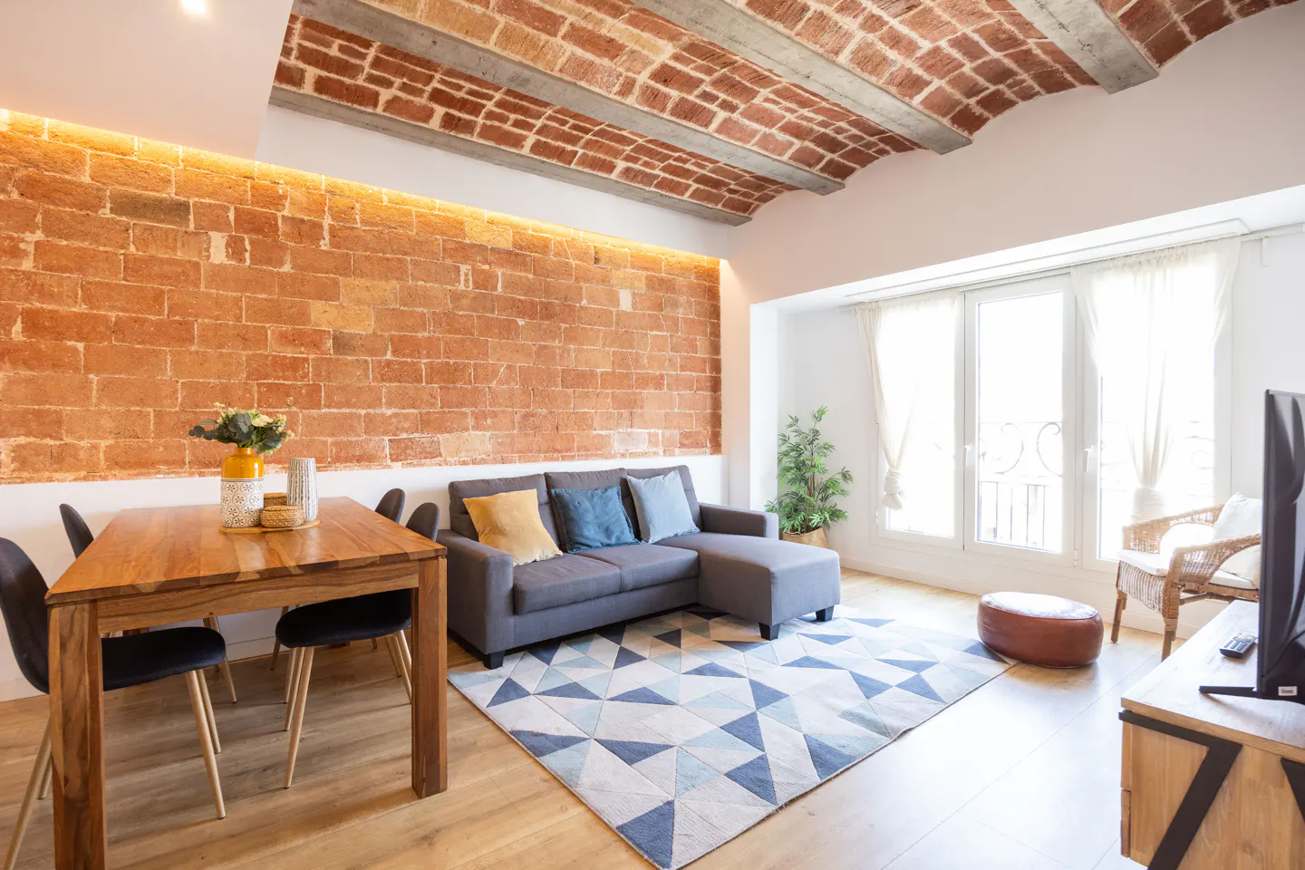 Bright living room with brick wall, wood floors, and a blue and white geometric rug. A gray sofa and wood table with chairs are visible.